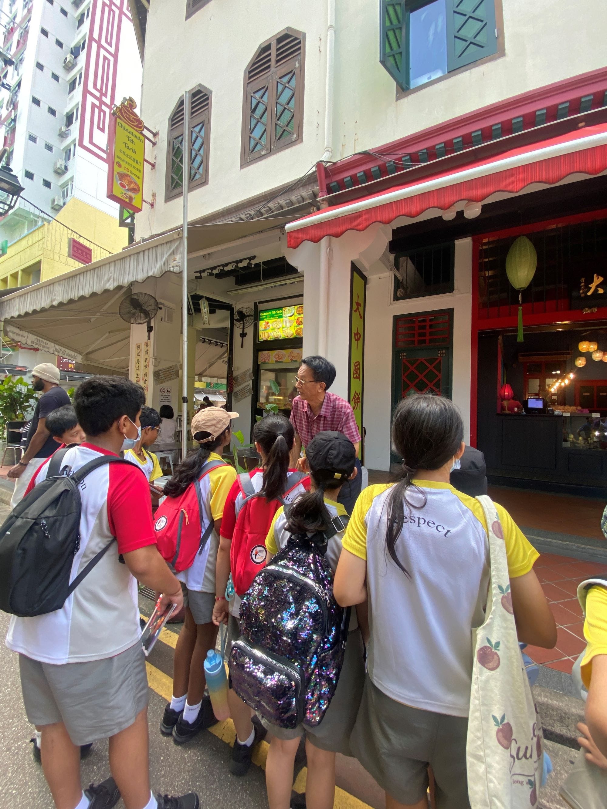 A group of school children with backpacks listen to an adult man outside colorful shophouses in Chinatown.